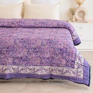 May include: A lavender floral quilt with a cream border and navy blue trim is draped over a bed. The quilt features a repeating pattern of flowers and leaves in shades of lavender and blue. Two cream-coloured pillows and two white pillows are visible.