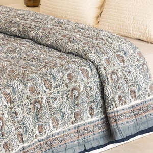 May include: A gray and white floral patterned quilt with a brown border. The quilt is folded on a bed with white pillows.