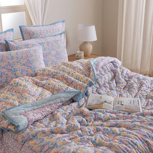 Op de afbeelding: Een bed is opgemaakt met een bloemmotief quilt, kussenslopen en een decoratieve rand. De quilt en kussenslopen hebben een herhalend patroon van gele, roze en blauwe bloemen op een lichtblauwe achtergrond. Een boek ligt open op het bed.
