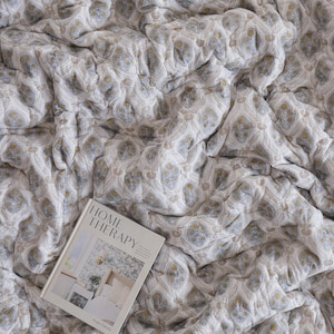 May include: A quilted textile featuring a repeating damask-style pattern in muted blue and mustard yellow on a white background. A hardcover book titled HOME THERAPY with a photograph of a bedroom interior on the cover rests on the fabric surface.