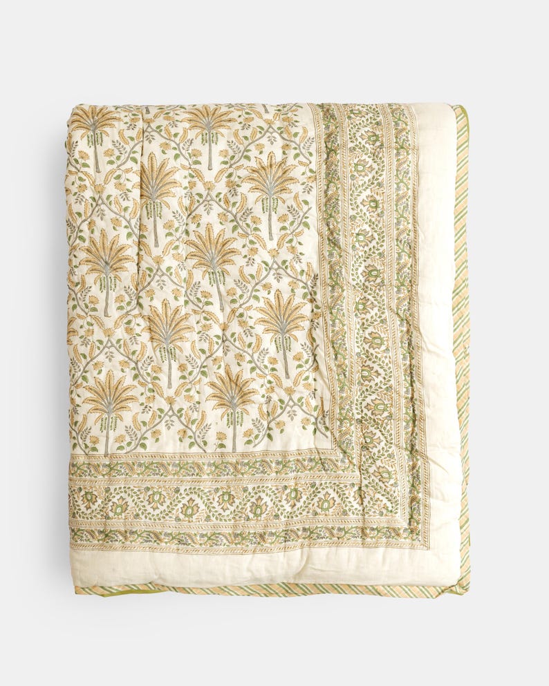May include: A folded, cream-colored quilt with a repeating pattern of stylized palm trees in shades of gold and green. The border features a detailed floral design in green and cream, with a striped edge.