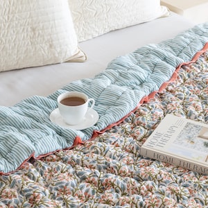 May include: A bed with a floral patterned quilt and a striped blue and white quilt. A white teacup and saucer sit on the striped quilt. A book titled "HOME THERAPY" rests on the floral quilt. White pillows are in the background.