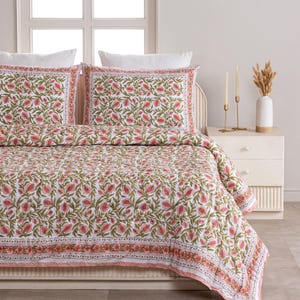 May include: A white and pink floral patterned quilt set with two matching pillowcases. The bedding features a repeating design of pink flowers and green leaves on a white background. The quilt and pillowcases have a decorative border.