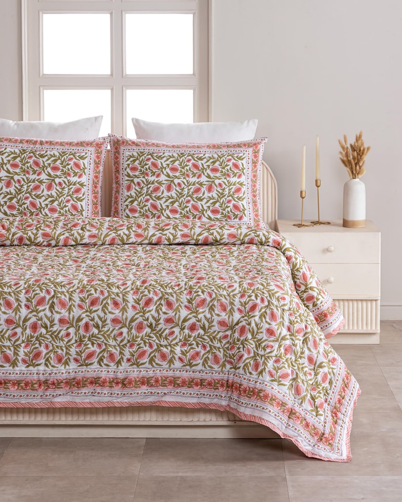 May include: A white and pink floral patterned quilt set with two matching pillowcases. The bedding features a repeating design of pink flowers and green leaves on a white background. The quilt and pillowcases have a decorative border.