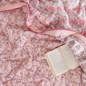 May include: A soft, quilted cotton blanket featuring a repeating floral block print in shades of dusty pink, white, and muted brown. The blanket has a striped red and white fabric trim along the edge and is partially folded over an open book.