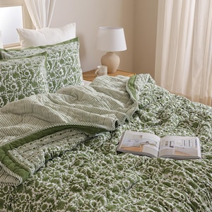 Könnte beinhalten: Ein Bett mit einer grün-weißen Steppdecke mit Blumenmuster und passenden Kissenbezügen. Eine gestreifte Steppdecke ist am Fußende des Bettes gefaltet. Ein offenes Buch liegt auf der Steppdecke. Eine weiße Lampe und eine Tasse stehen auf einem Nachttisch aus Holz.