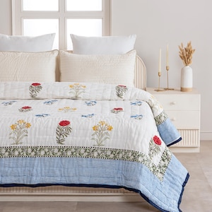 Puede incluir: Un edred&oacute;n blanco con un estampado floral con flores rojas, amarillas y azules. El edred&oacute;n tiene un borde a rayas azules y se muestra en una cama con almohadas blancas. El juego de cama incluye un dise&ntilde;o floral a juego.