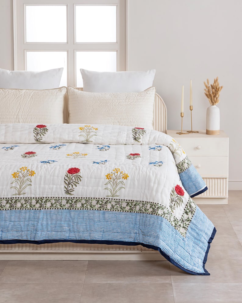 Puede incluir: Un edred&oacute;n blanco con un estampado floral con flores rojas, amarillas y azules. El edred&oacute;n tiene un borde a rayas azules y se muestra en una cama con almohadas blancas. El juego de cama incluye un dise&ntilde;o floral a juego.