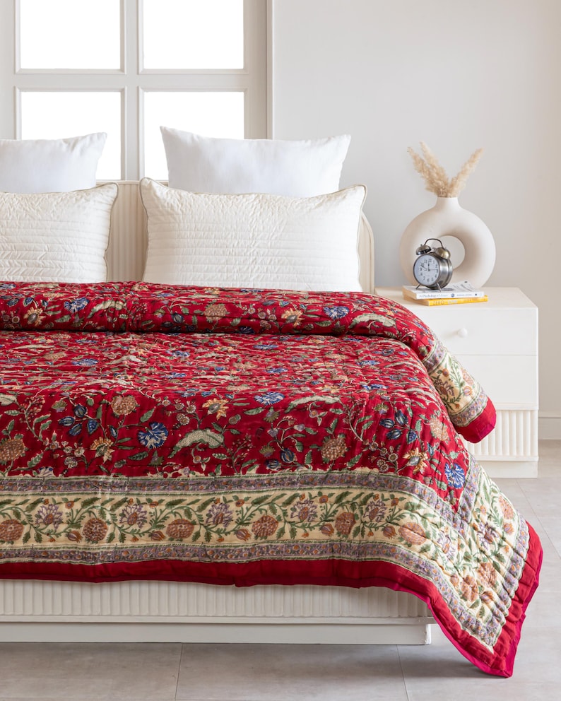 May include: A red floral quilt with a cream border and a floral pattern. The quilt is on a white bed with white pillows.