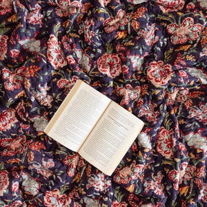 K&ouml;nnte beinhalten: Ein offenes Buch liegt auf einer dunkelblauen und roten, mit Blumenmustern bedruckten Steppdecke. Das Buch ist auf einer Seite mit Text aufgeschlagen.