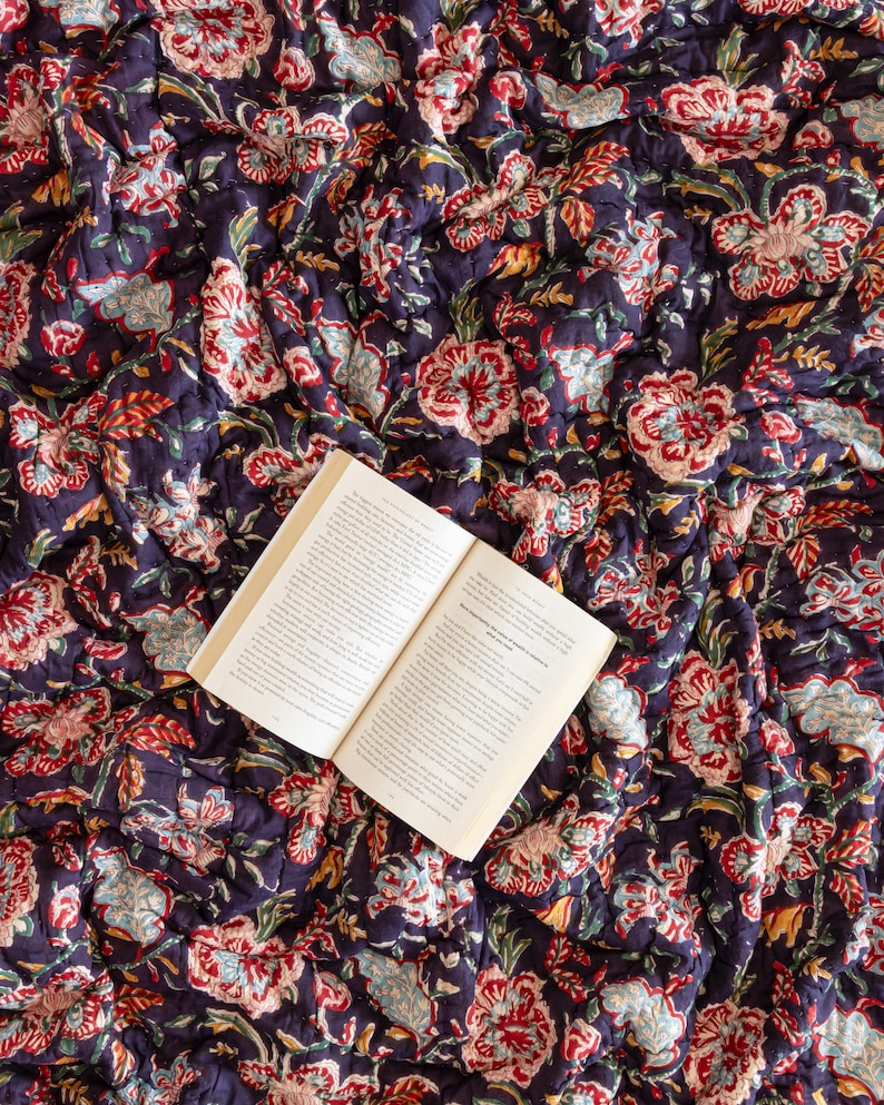 K&ouml;nnte beinhalten: Ein offenes Buch liegt auf einer dunkelblauen und roten, mit Blumenmustern bedruckten Steppdecke. Das Buch ist auf einer Seite mit Text aufgeschlagen.