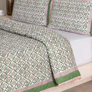 May include: A white and green floral patterned quilt and pillow set. The bedding features a repeating diamond pattern with pink flowers and green leaves. The edges of the quilt and pillows are trimmed with a solid green border.