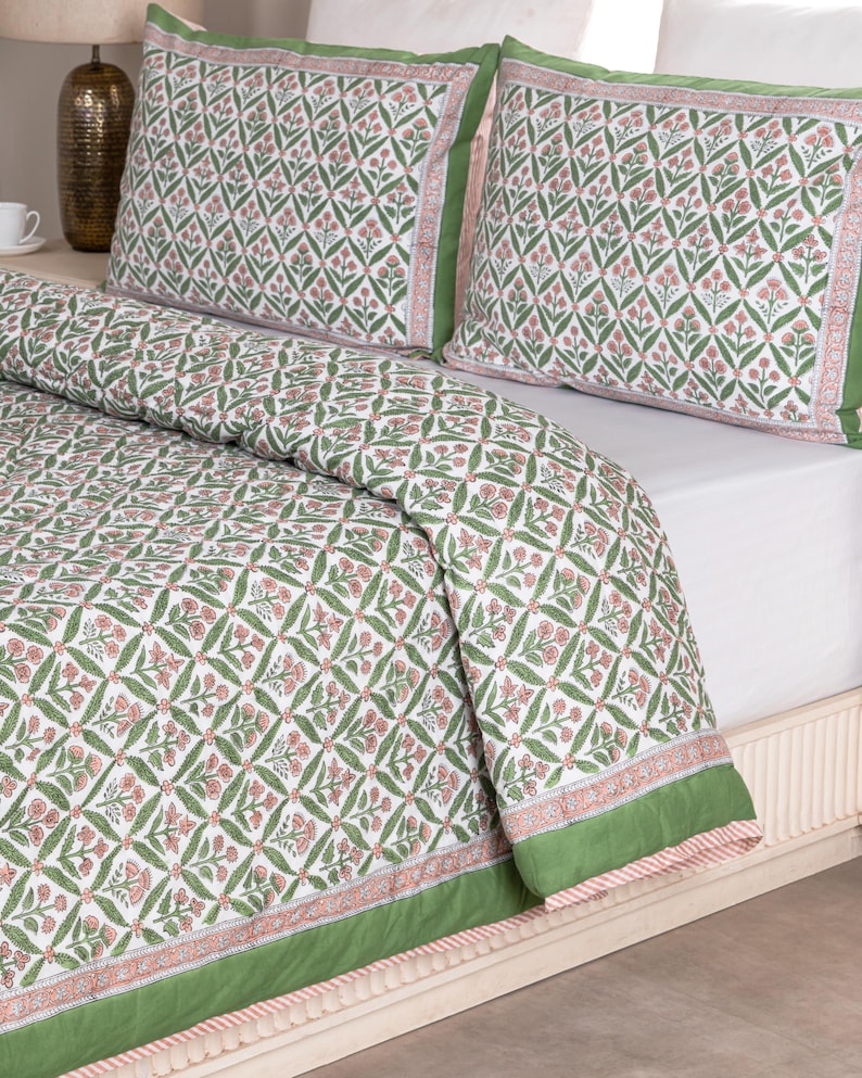 May include: A white and green floral patterned quilt and pillow set. The bedding features a repeating diamond pattern with pink flowers and green leaves. The edges of the quilt and pillows are trimmed with a solid green border.