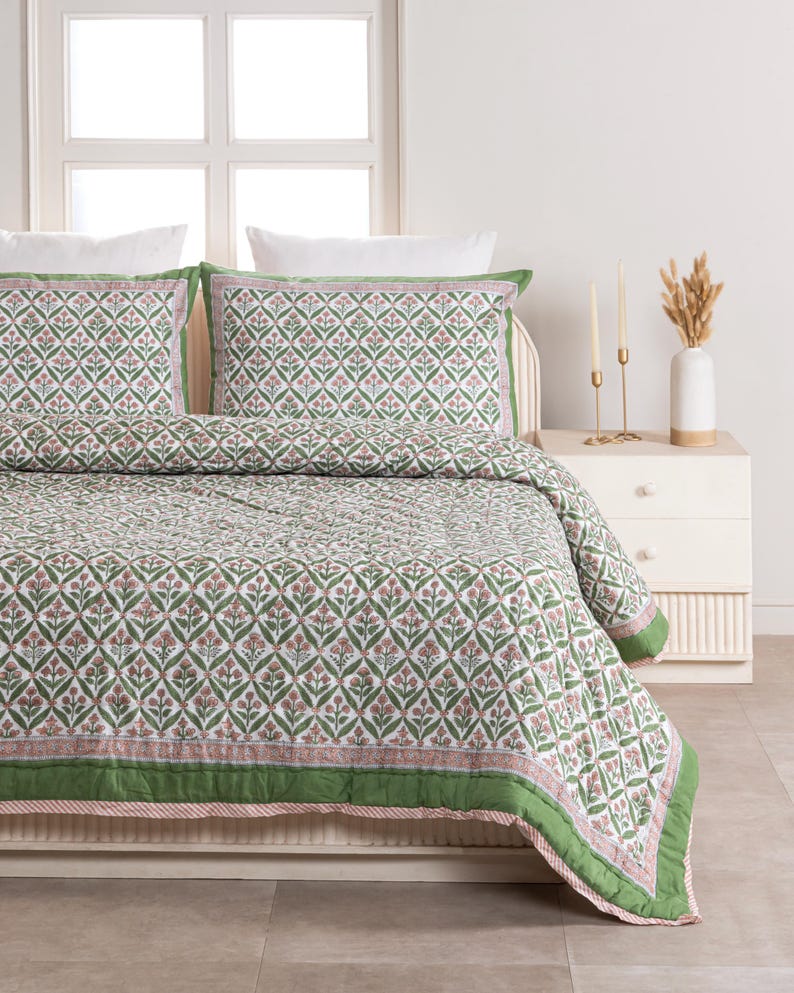 May include: A bed with a floral patterned quilt and matching pillowcases. The quilt features a repeating pattern of green leaves and pink flowers on a white background, with a green border. The pillowcases have a similar design.