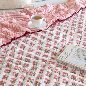 Puede incluir: Una cama está cubierta con un edredón blanco con estampado floral y un edredón a rayas rojas y blancas. Una taza y un platillo blancos están sobre el edredón a rayas. Un libro titulado "HOME THERAPY" descansa sobre el edredón floral.