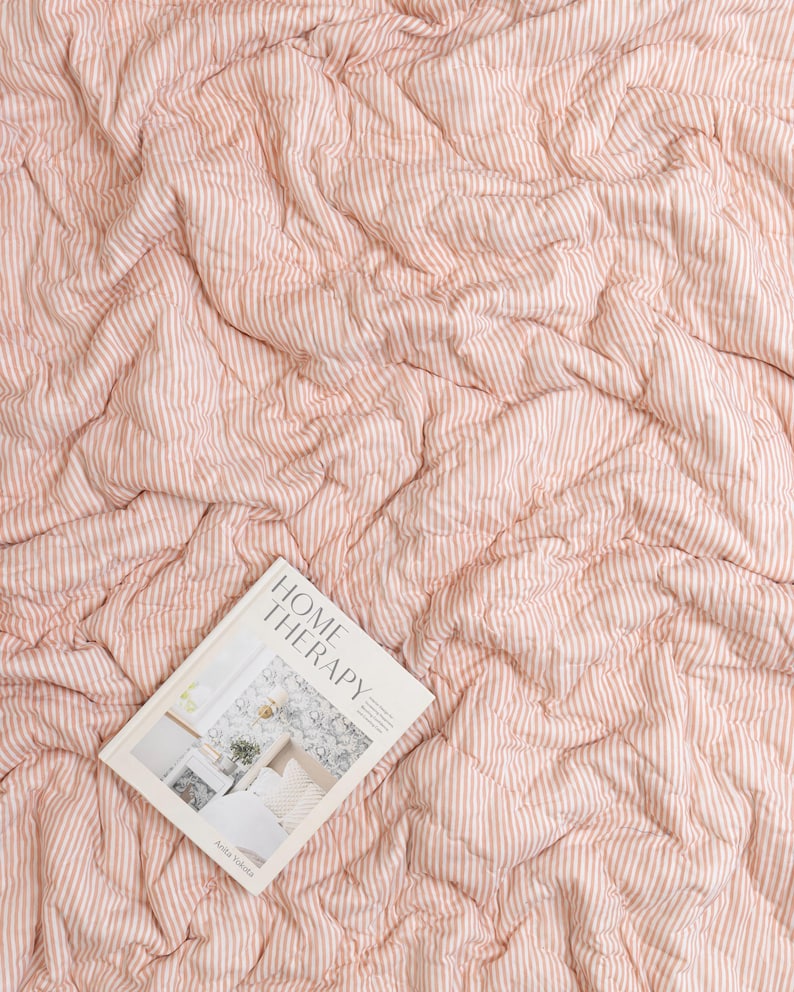 May include: A quilted, peach-colored blanket with vertical white stripes. The blanket is rumpled, creating a textured appearance. A book titled "HOME THERAPY" is lying on top of the blanket.