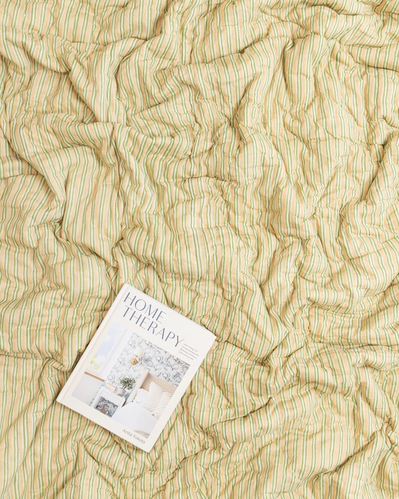 May include: A cream-colored, striped quilt with vertical green stripes. A book titled "HOME THERAPY" is lying on top of the quilt. The book cover features a bedroom scene.