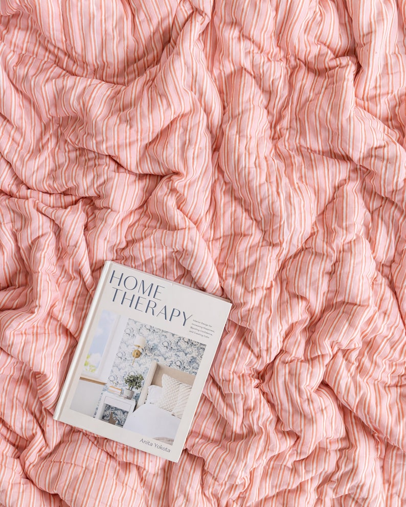May include: A pink and white striped quilt with a book titled "HOME THERAPY" by Anita Yokota. The quilt has a textured, quilted design. The book cover features a bedroom scene.