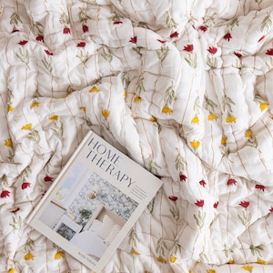 Puede incluir: Manta acolchada blanca con un estampado floral en amarillo y rojo. La manta tiene l&iacute;neas verticales cosidas. Un libro titulado "HOME THERAPY" est&aacute; sobre la manta.