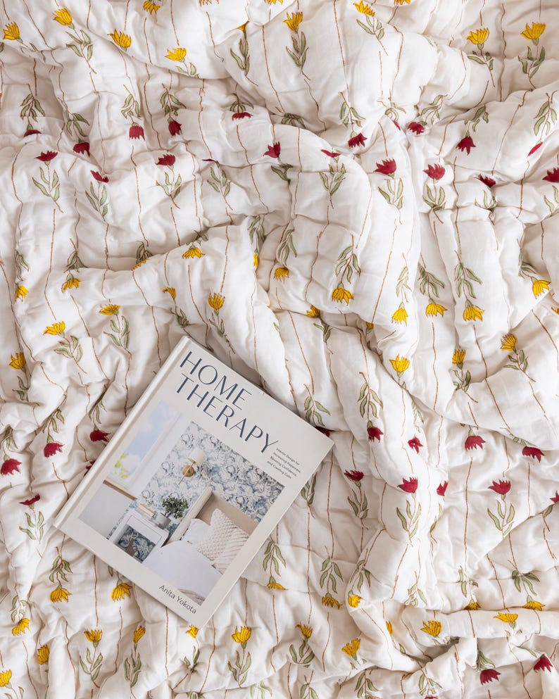 Puede incluir: Manta acolchada blanca con un estampado floral en amarillo y rojo. La manta tiene l&iacute;neas verticales cosidas. Un libro titulado "HOME THERAPY" est&aacute; sobre la manta.