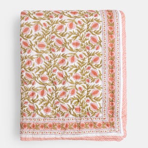 May include: A folded white quilt with a repeating pattern of pink pomegranates and green leaves. The quilt has a pink border with a floral design and a striped edge. The overall design is a floral theme.