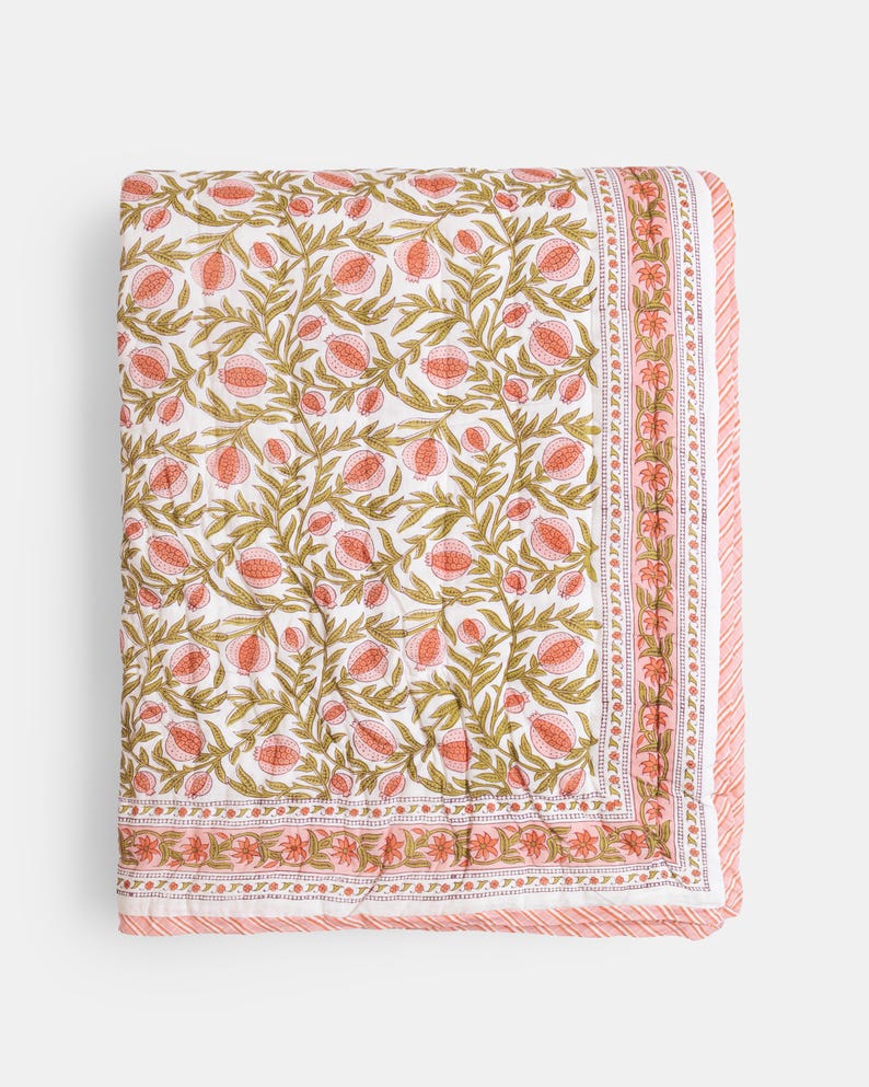 May include: A folded white quilt with a repeating pattern of pink pomegranates and green leaves. The quilt has a pink border with a floral design and a striped edge. The overall design is a floral theme.