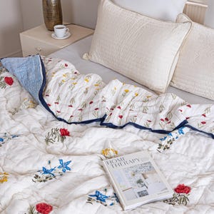 Puede incluir: Colcha acolchada blanca con un estampado floral en rojo, amarillo y azul. La colcha tiene un borde azul marino y se combina con almohadas blancas. Un libro titulado "HOME THERAPY" descansa sobre la colcha.