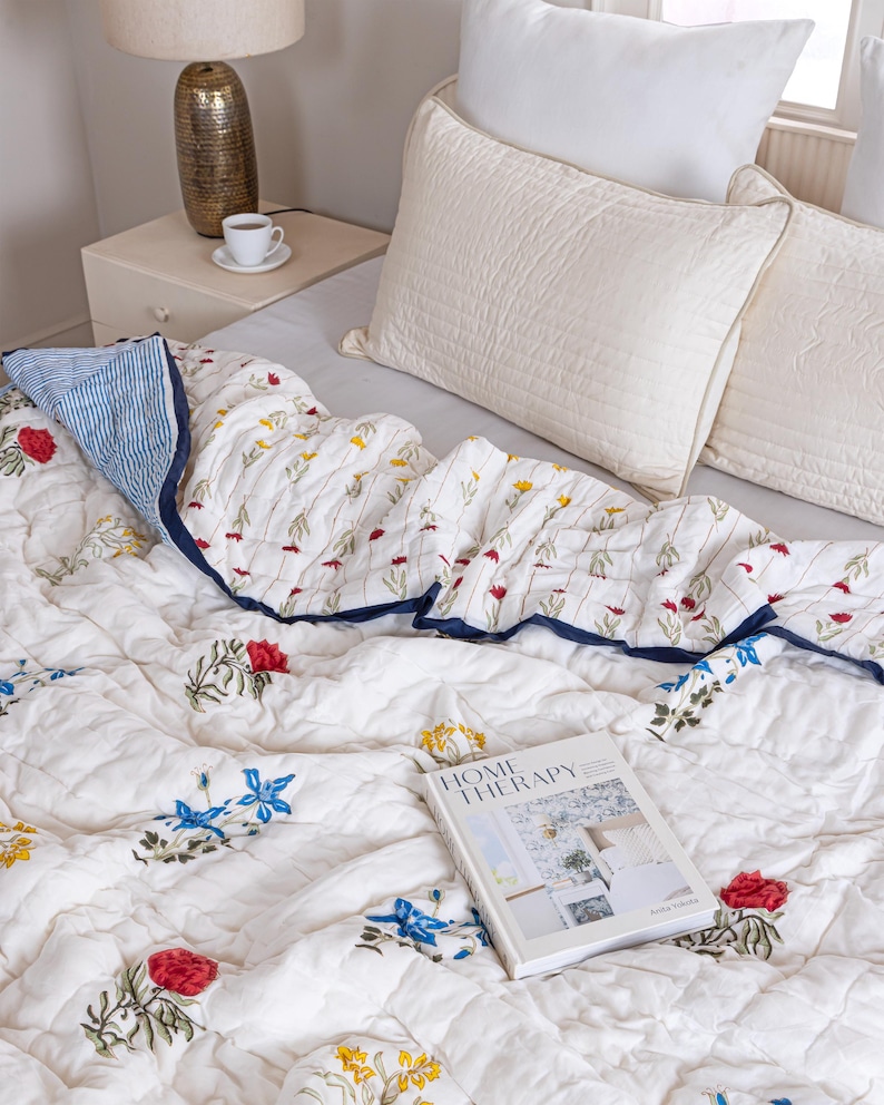 Puede incluir: Colcha acolchada blanca con un estampado floral en rojo, amarillo y azul. La colcha tiene un borde azul marino y se combina con almohadas blancas. Un libro titulado "HOME THERAPY" descansa sobre la colcha.