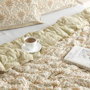 May include: A bed is made with a floral patterned pillowcase and a quilted comforter. The comforter has a striped section and a floral section. A white teacup and saucer sit on the comforter, and a book titled "HOME THERAPY" is also on the bed.