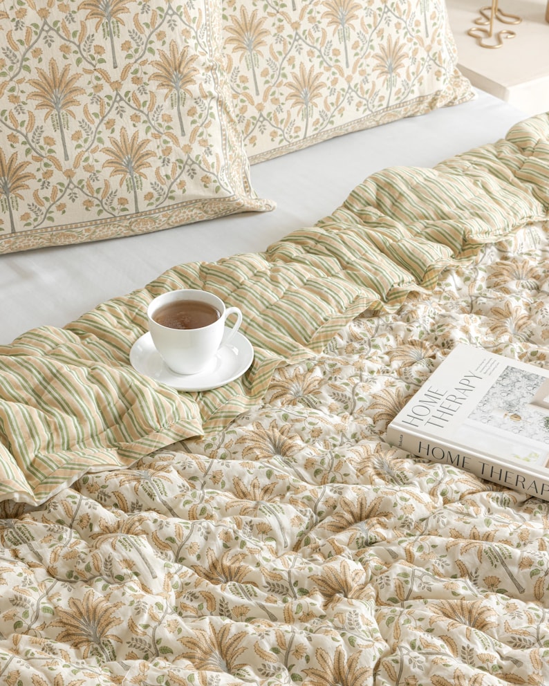 May include: A bed is made with a floral patterned pillowcase and a quilted comforter. The comforter has a striped section and a floral section. A white teacup and saucer sit on the comforter, and a book titled "HOME THERAPY" is also on the bed.