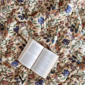 May include: A cream-colored quilt with a floral pattern in shades of blue, green, and orange. The quilt is folded and a book is lying on top of it.