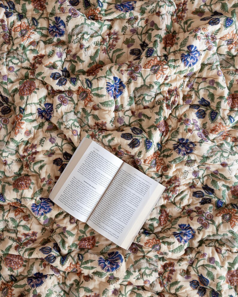 May include: A cream-colored quilt with a floral pattern in shades of blue, green, and orange. The quilt is folded and a book is lying on top of it.