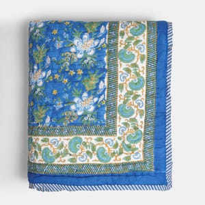 Könnte beinhalten: Eine gefaltete blaue Steppdecke mit floralem Muster. Die Steppdecke zeigt große weiße Blumen mit blauen Akzenten auf blauem Hintergrund. Ein Rand mit grünen und blauen Blumenmustern und ein gestreifter Rand vervollständigen das Design.