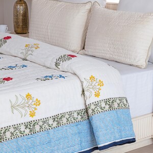 Puede incluir: Colcha blanca con bordados florales en rojo, amarillo y azul. La colcha presenta un borde a rayas azules y blancas. Se ven dos almohadas blancas acolchadas en la cama. La colcha es probablemente de algod&oacute;n.