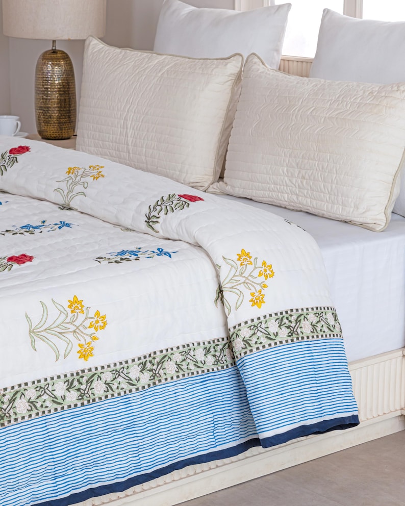 Puede incluir: Colcha blanca con bordados florales en rojo, amarillo y azul. La colcha presenta un borde a rayas azules y blancas. Se ven dos almohadas blancas acolchadas en la cama. La colcha es probablemente de algod&oacute;n.