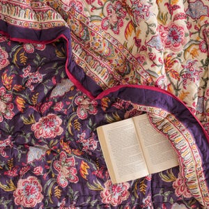 May include: A reversible quilted blanket featuring a floral pattern in shades of deep purple, cream, pink, and yellow, with a dark purple border and a pink trim. An open book with printed text rests on the surface of the folded fabric.