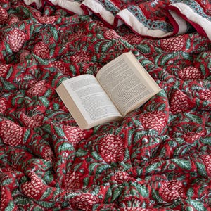 May include: A red and green patterned quilt with a book lying on top. The quilt has a floral design with a white background.