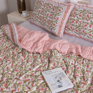 May include: A bed is made with a white sheet, two matching pillowcases, and a patterned quilt. The quilt and pillowcases feature a white background with a repeating pattern of pink flowers and green leaves. A book titled "HOME THERAPY" rests on the quilt.