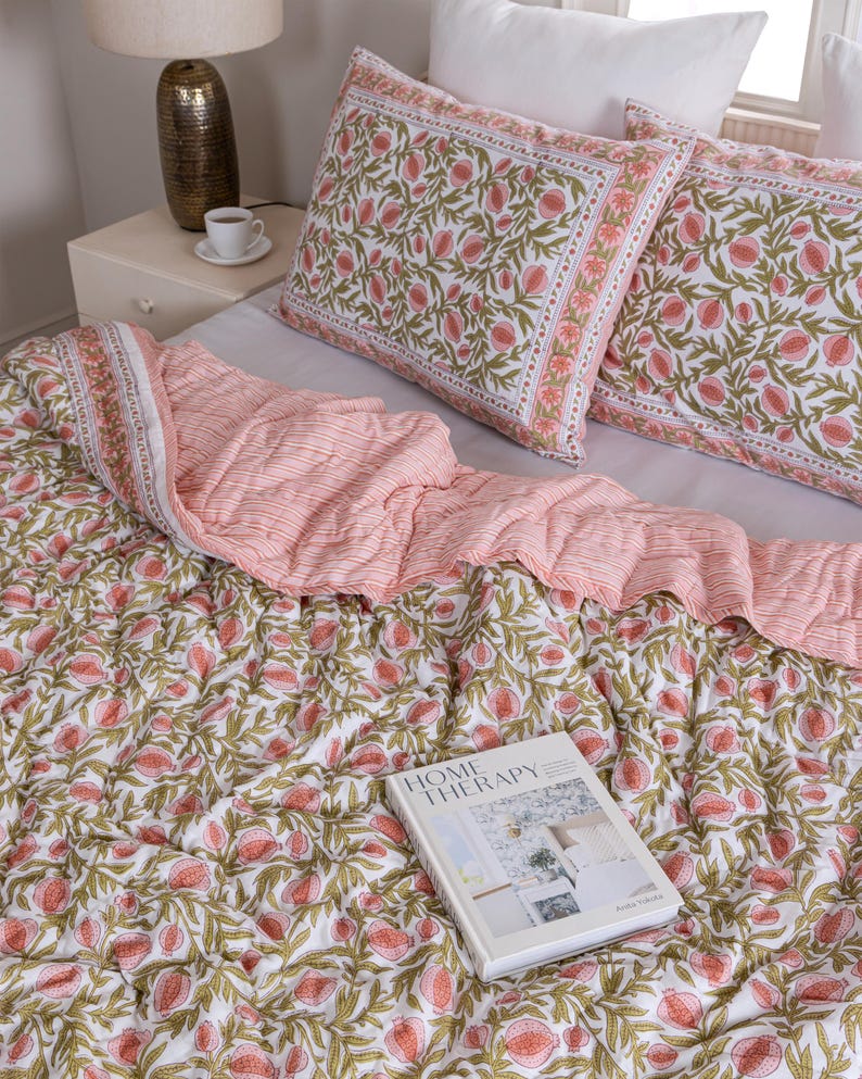 May include: A bed is made with a white sheet, two matching pillowcases, and a patterned quilt. The quilt and pillowcases feature a white background with a repeating pattern of pink flowers and green leaves. A book titled "HOME THERAPY" rests on the quilt.