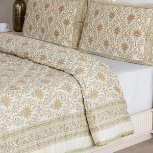 May include: A cream-colored bedding set featuring a floral pattern in shades of tan, green, and light blue. The set includes a quilt and two pillowcases. The quilt has a decorative border with a green and white striped trim.