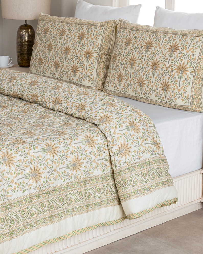 May include: A cream-colored bedding set featuring a floral pattern in shades of tan, green, and light blue. The set includes a quilt and two pillowcases. The quilt has a decorative border with a green and white striped trim.