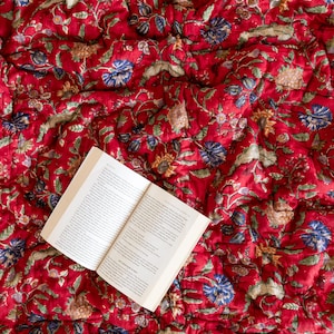 May include: An open book with white pages lies on a red and white floral patterned quilt. The quilt has a traditional Indian design.