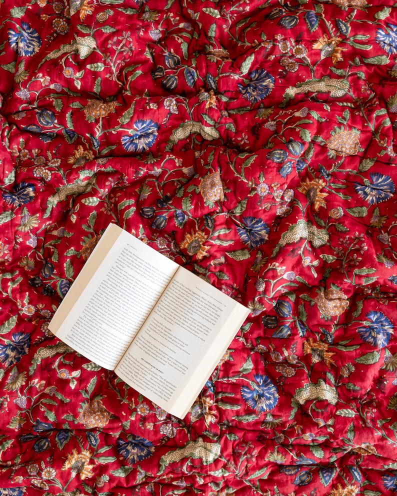 May include: An open book with white pages lies on a red and white floral patterned quilt. The quilt has a traditional Indian design.