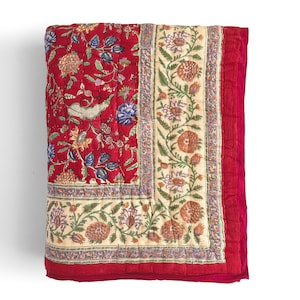 May include: A red and cream floral patterned quilt with a border of green and purple flowers. The quilt is folded in half and has a soft, quilted texture.