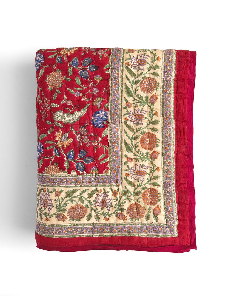 May include: A red and cream floral patterned quilt with a border of green and purple flowers. The quilt is folded in half and has a soft, quilted texture.
