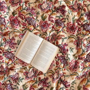 K&ouml;nnte beinhalten: Eine wei&szlig;e Baumwolldecke mit einem floralen Muster in Rot-, Lila- und Gr&uuml;nt&ouml;nen. Die Decke ist gefaltet und ein Buch liegt darauf.