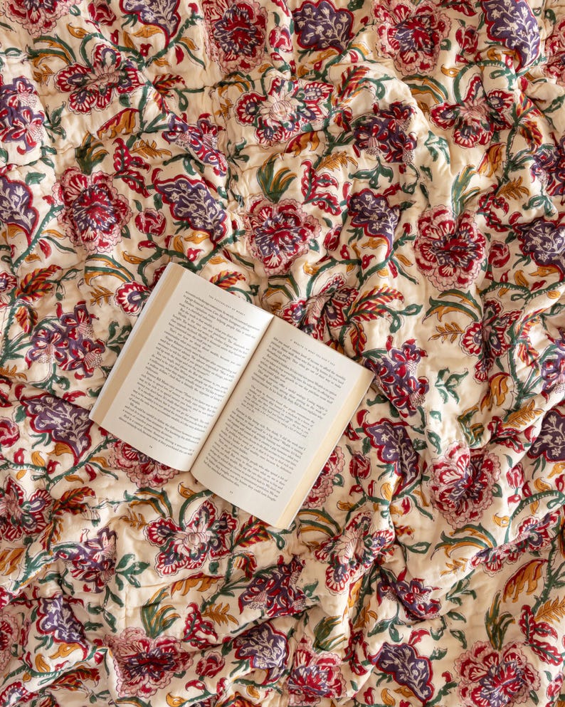 K&ouml;nnte beinhalten: Eine wei&szlig;e Baumwolldecke mit einem floralen Muster in Rot-, Lila- und Gr&uuml;nt&ouml;nen. Die Decke ist gefaltet und ein Buch liegt darauf.