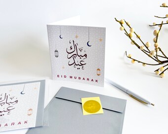 May include: A white card with a gray and white patterned background. The card has Arabic calligraphy in brown and red, and the text "Eid Mubarak" in red. The card is sitting on a white surface with a gray envelope and a white pen.