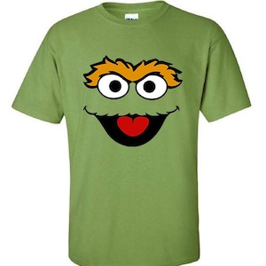 May include: A green t-shirt featuring a cartoon face with large white eyes, orange eyebrows, a black mouth, and a red tongue. The design is centered on the front of the shirt. The shirt is made of a soft, comfortable material.