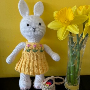 Pode incluir: Um coelho de croch&ecirc; branco vestindo um vestido amarelo com flores bordadas em rosa e verde. O coelho est&aacute; segurando uma pequena cesta de croch&ecirc; marrom com ovos de P&aacute;scoa coloridos dentro. O coelho est&aacute; em p&eacute; na frente de um vaso com narcisos amarelos.